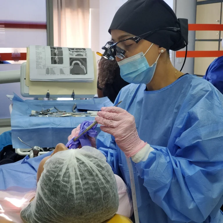 Female oral surgeon wearing surgical loupes and a blue gown placing a dental implant while reviewing a CT scan bone density scan