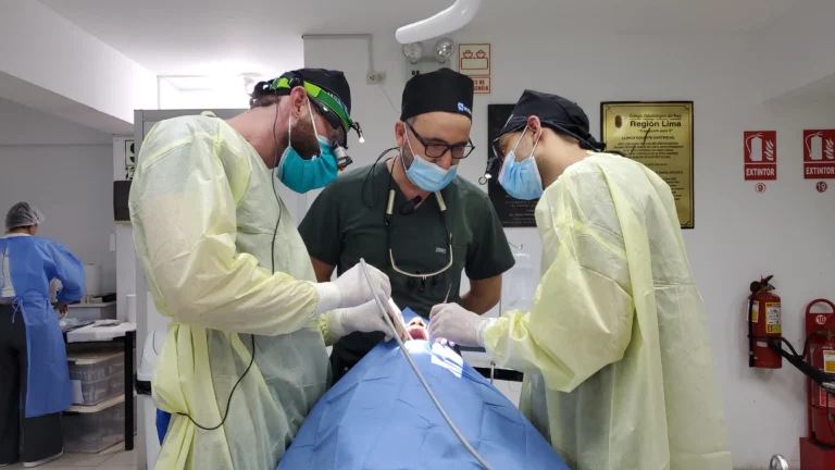 Three dentists in surgical attire performing a live dental implant procedure on a patient in a clinic with fire extinguishers and a plaque on the wall.